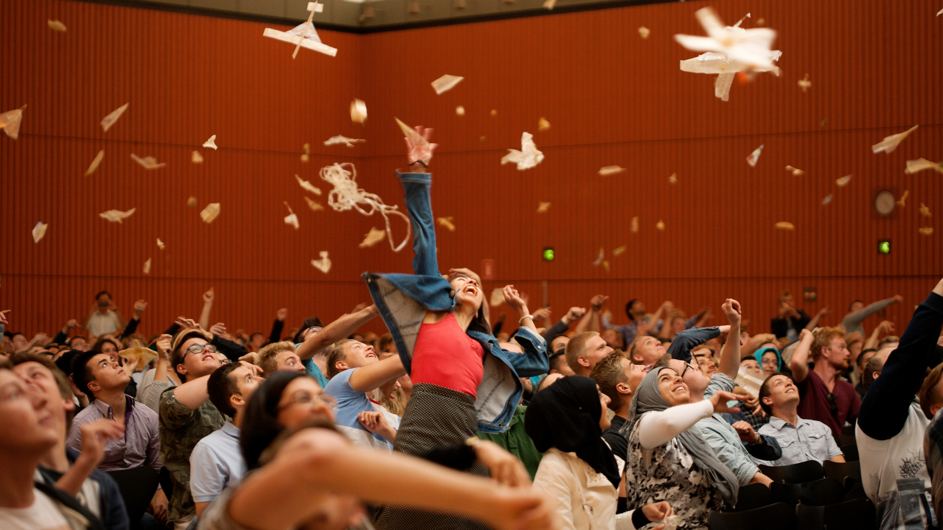 En gruppe glade studerende i et auditorium kaster papirflyvere op i luften med stor entusiasme